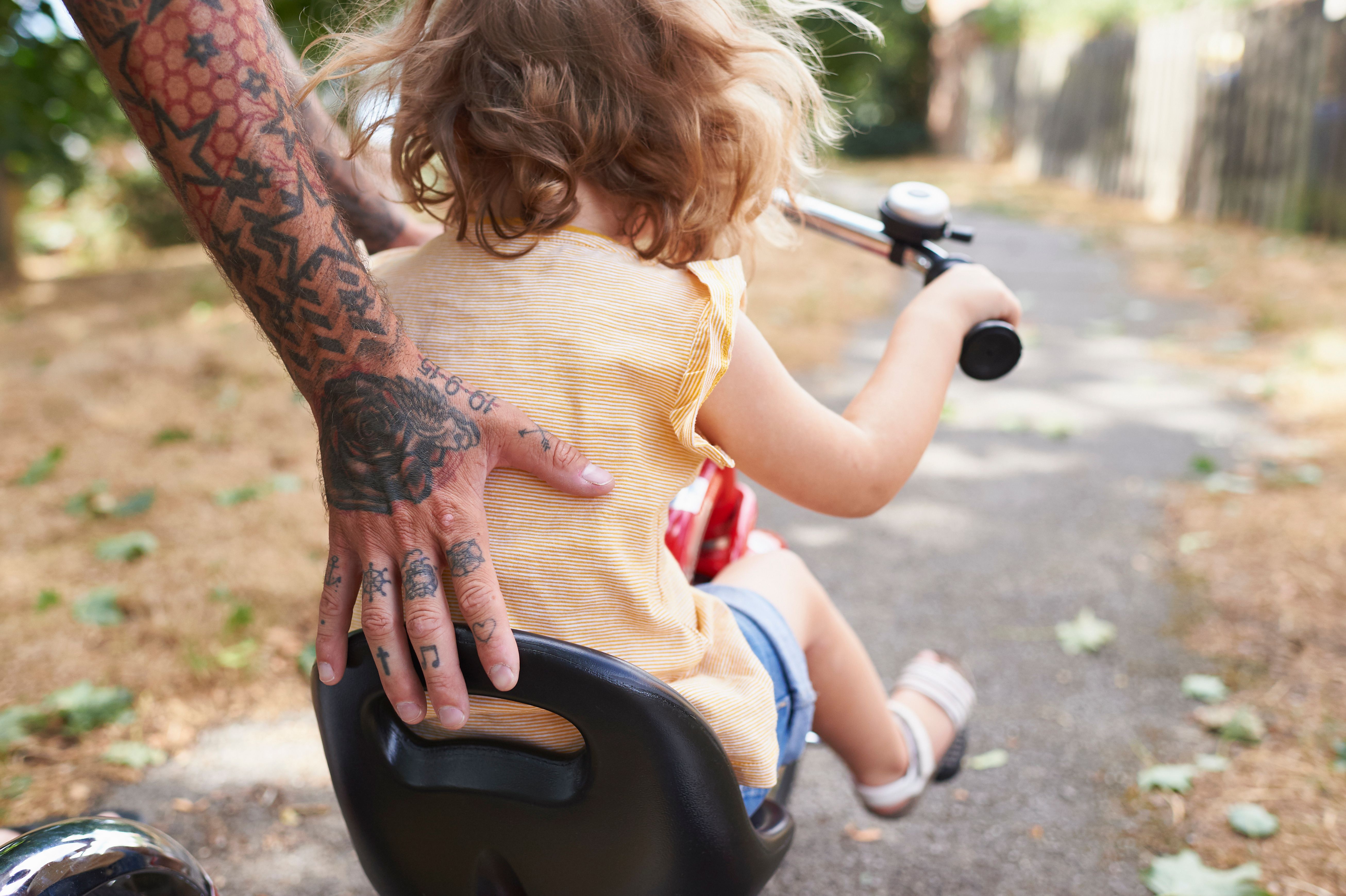 Child riding on a bicycle seat with an adult nearby on a tree-lined path.