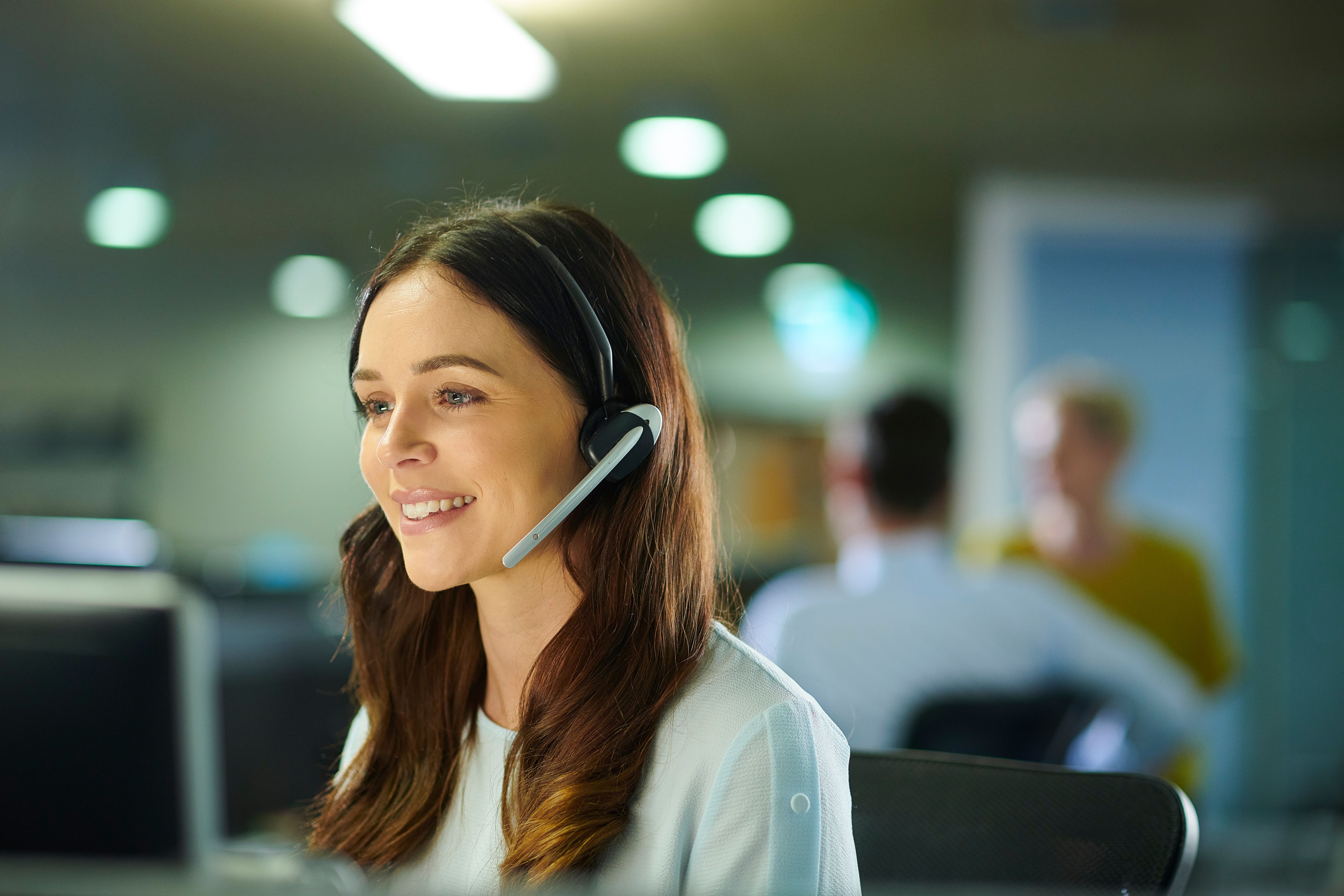 A woman wearing a headset, smiling while working at a computer in an office.
