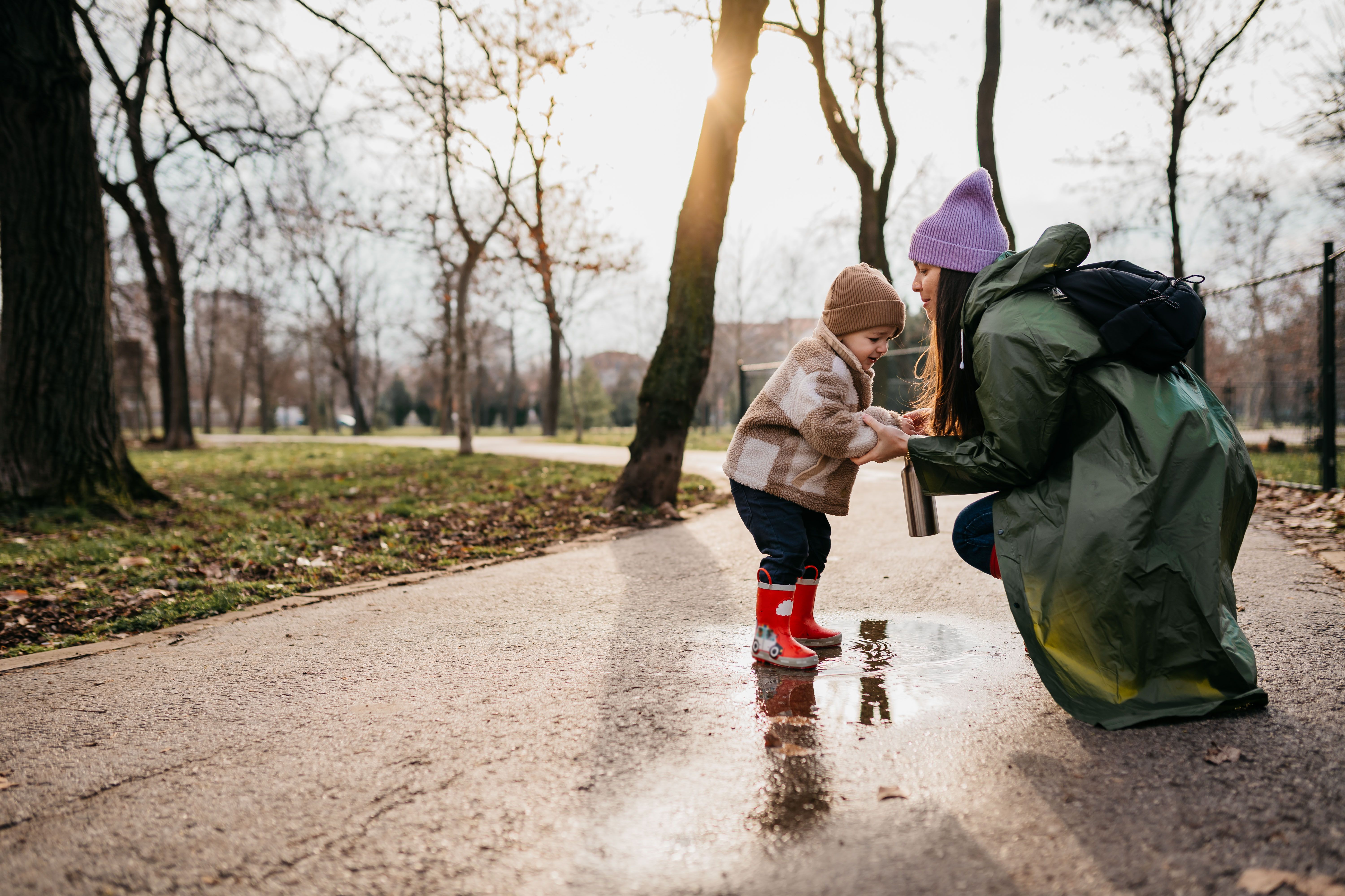 A women crouches to play with a toddler on a sunny park path surrounded by trees