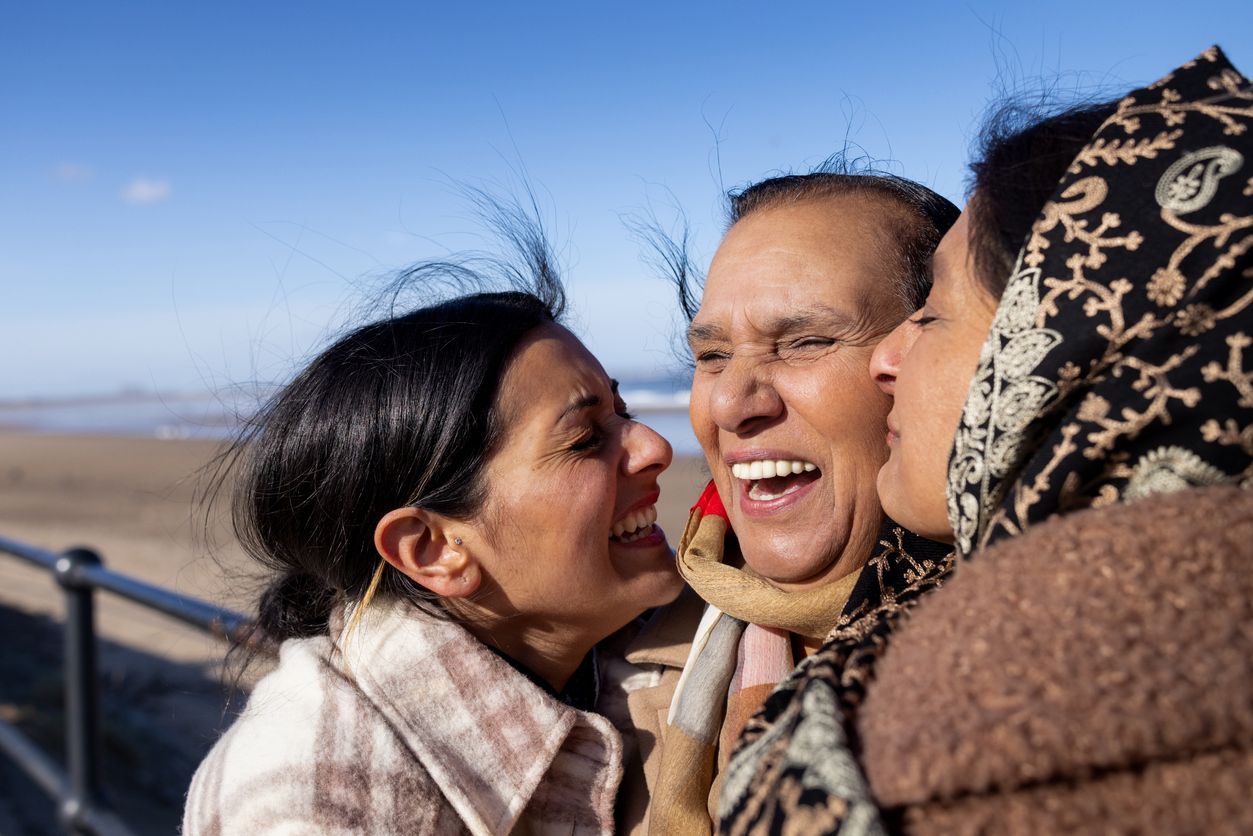 Grandmother, mother, and adult granddaughter laugh together on a sunny beach