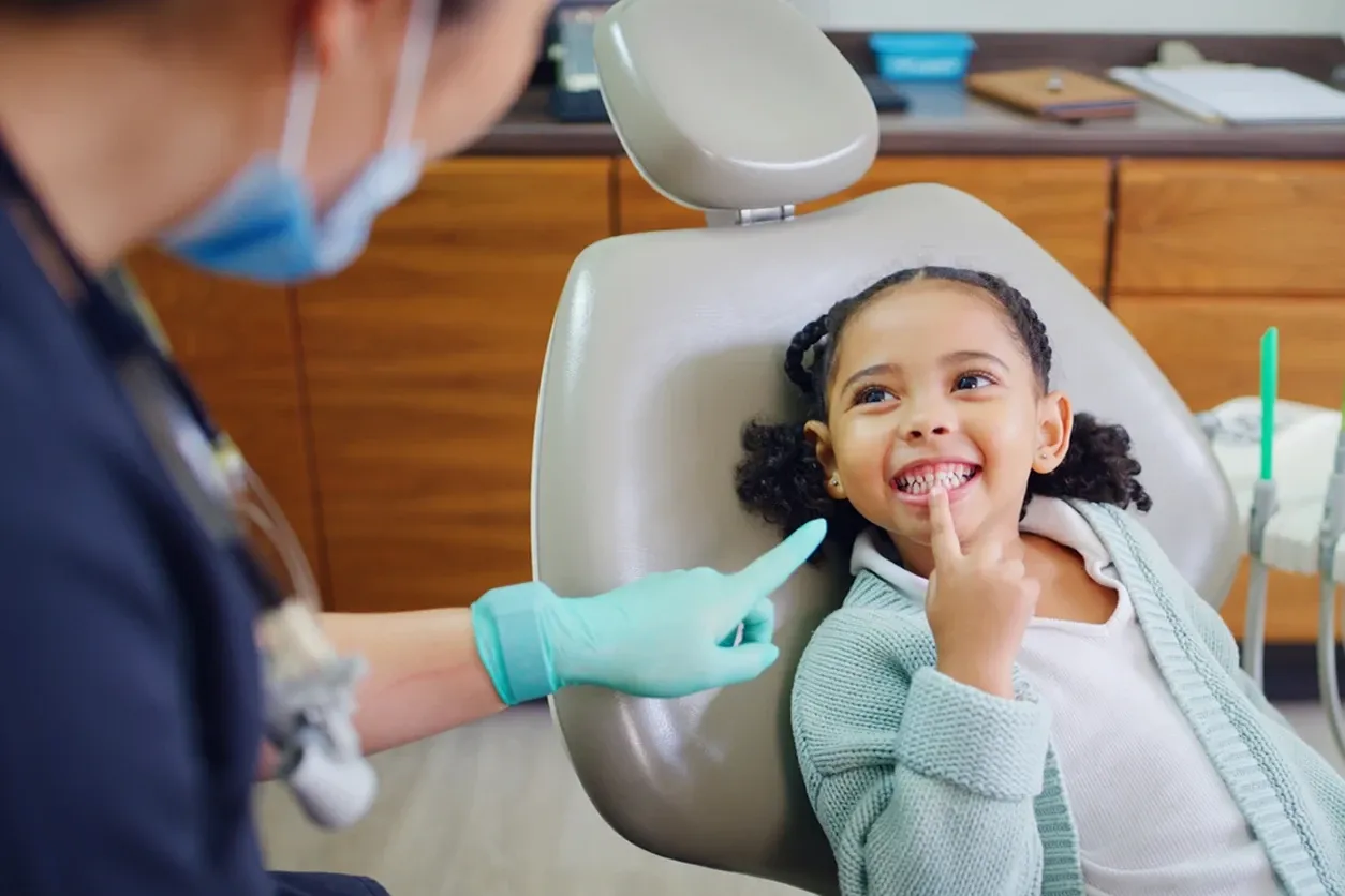A young girl with braided hair sits in a dental chair, smiling and pointing to her teeth while a dentist in a mask and gloves gestures toward her.