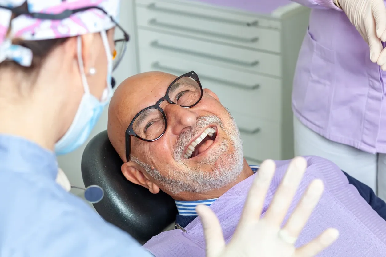 A smiling elderly man with glasses sits in a dental chair, looking up at a dentist wearing a mask and cap, with a dental assistant standing nearby.