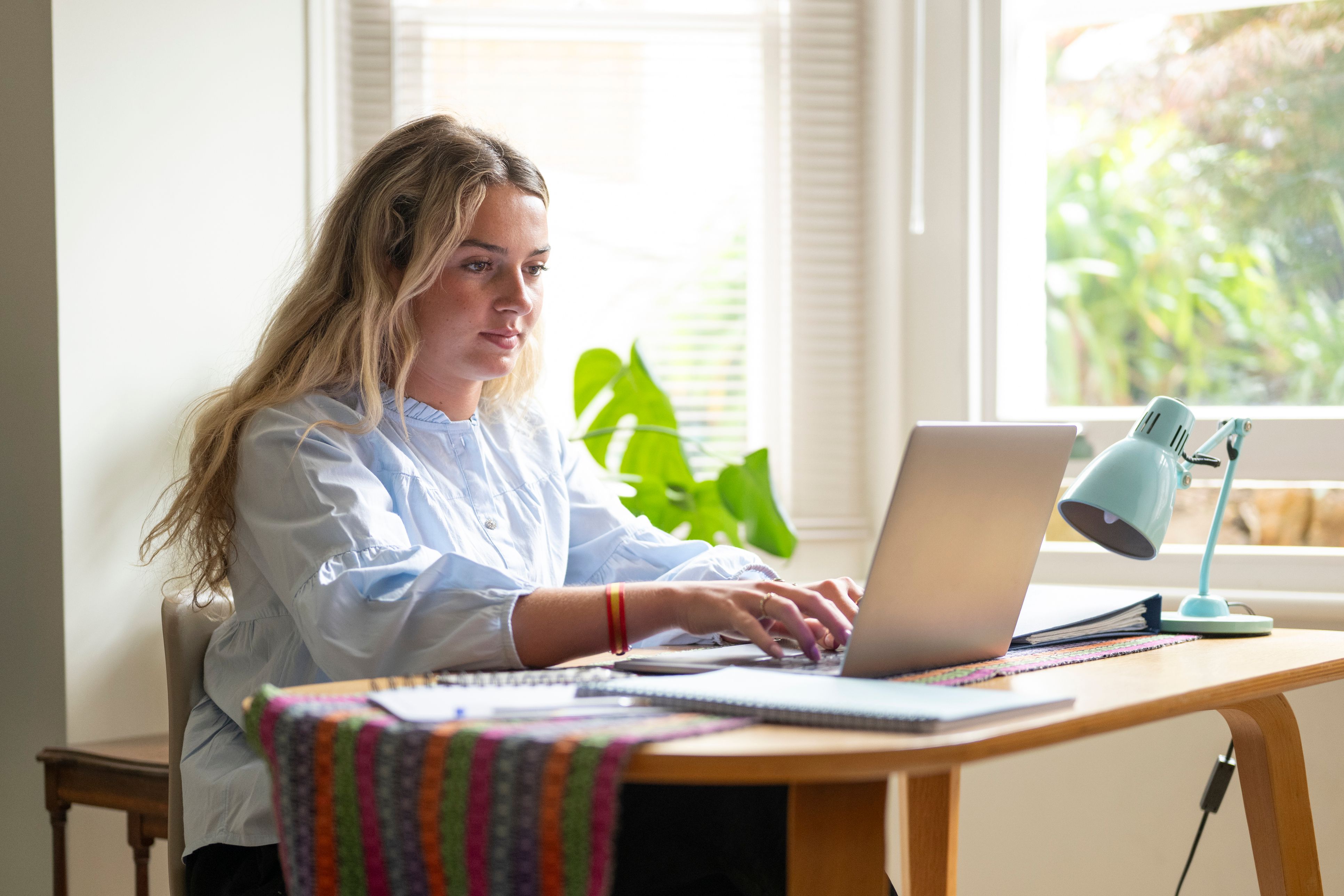 A woman sitting in her home at a desk by the window, working on her laptop with notebooks around her.