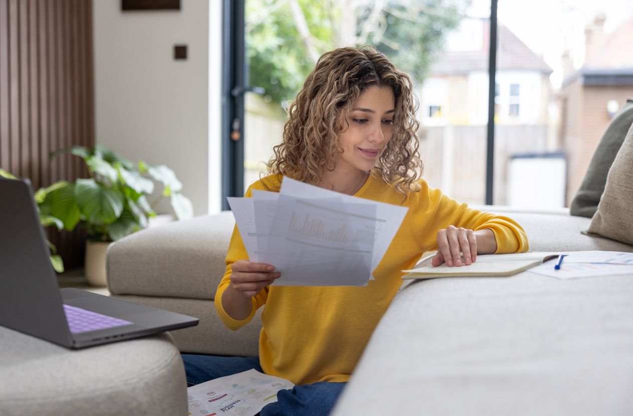 Women sitting on the floor, with a laptop on the sofa and reading letters/documents. 